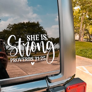 May include: White vinyl decal applied to a vehicle window. The decal reads "SHE IS Strong" with "PROVERBS 31:25" below, and a small heart. The background shows a reflection of trees and sky.