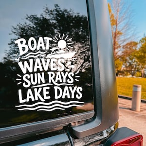 May include: A white vinyl decal applied to a vehicle window featuring a graphic of a boat, a sun, and wavy lines. The text reads: BOAT WAVES SUN RAYS LAKE DAYS. The design is composed of bold, stylized lettering and simple line art illustrations.