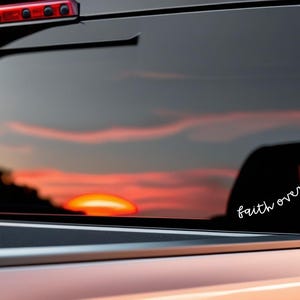 May include: A vehicle's rear window with a sunset reflection. A white decal on the glass reads "faith over fear" in cursive with a cross. The vehicle's body is a light orange color.