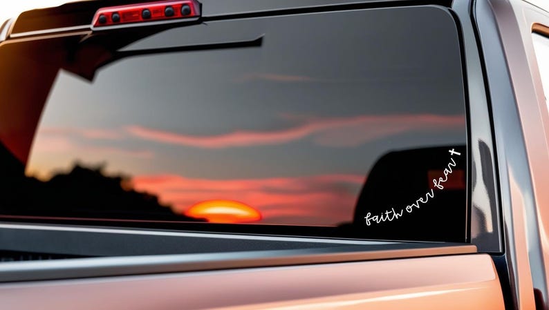 May include: A vehicle's rear window with a sunset reflection. A white decal on the glass reads "faith over fear" in cursive with a cross. The vehicle's body is a light orange color.