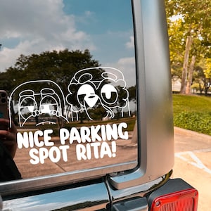 May include: A white vinyl decal on a car window features two cartoon characters with the text "NICE PARKING SPOT RITA!". The decal is applied to the rear window of a vehicle, with a view of trees and sky reflected in the glass.