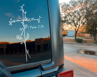 May include: White vinyl decal of a cross with floral accents, applied to a vehicle window. The text "I Peter 2:24" is below the cross. The vehicle is black, with a red taillight visible. The background includes trees and a bright sky.