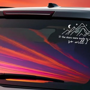 May include: A white car with a sunset reflected in the back window. The window has a white vinyl decal with a mountain range, stars, and a crescent moon. The decal also includes the text "If the stars were made to worship, so will I."