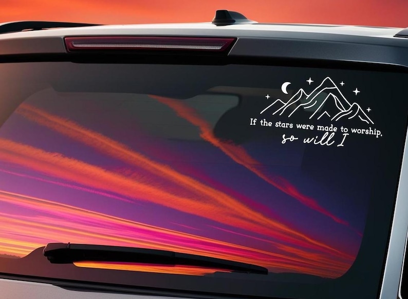 May include: A white car with a sunset reflected in the back window. The window has a white vinyl decal with a mountain range, stars, and a crescent moon. The decal also includes the text "If the stars were made to worship, so will I."