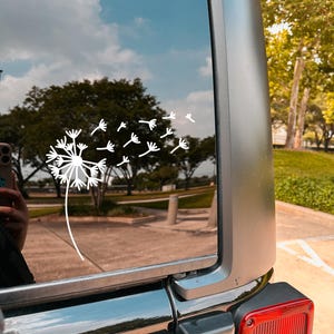 May include: A white dandelion sticker on the back window of a black car. The dandelion is blowing in the wind, with its seeds flying away.