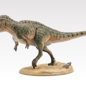 Puede incluir: Figura detallada de dinosaurio, probablemente un Allosaurus, en una pose dinámica. La figura es principalmente verde y marrón con detalles en naranja, sobre una base beige. La boca del dinosaurio está abierta y su cola se extiende hacia atrás.
