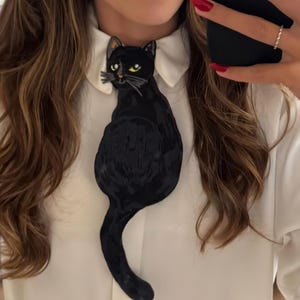 Peut inclure: Une broche noire en forme de chat avec des yeux jaunes et des moustaches est épinglée sur une chemise blanche à col. La broche est faite d'un matériau texturé et a une longue queue. Le mot "Wrapistry" est en bas de l'image.