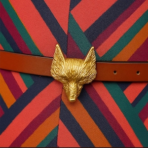 Könnte beinhalten: Nahaufnahme eines braunen Ledergürtels mit einer goldfarbenen Fuchskopf-Schnalle. Der Gürtel befindet sich vor einem Stoff mit roten, grünen und marineblauen Streifen. Das Wort "Wrapistry" steht unten.