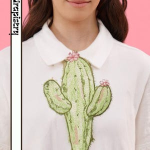 Peut inclure: Polo blanc avec un motif de cactus brodé en vert et rose. Le cactus a deux bras latéraux et une petite fleur rose au sommet. Le chemisier a un col classique et des manches courtes.