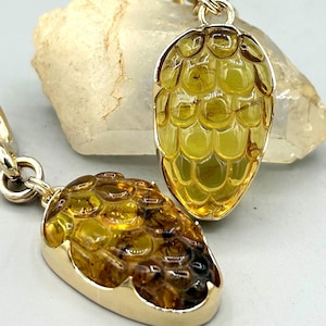 May include: Two gold-toned pendants shaped like pine cones. The pendants have a textured, amber-colored center and a gold border. The pendants are on a gold chain. The background includes a light-colored stone.