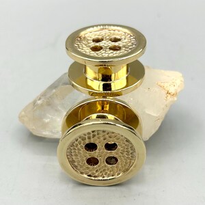 May include: Two gold-toned metal buttons with a smooth, polished outer rim and a textured, hammered centre surface. Each button features four central holes arranged in a square pattern, reflecting a bright, metallic finish throughout the design.