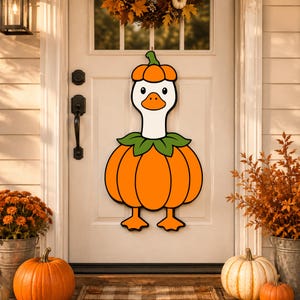 Può includere: Una decorazione da porta a forma di oca bianca che indossa un colletto verde e un corpo a forma di zucca arancione, con un piccolo cappello a zucca arancione sulla testa. L'oca ha un becco arancione brillante e zampe palmate arancioni, contornate di nero.