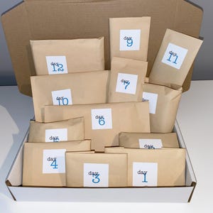 May include: An open cardboard box filled with brown paper packages. Each package has a white label with the word "day" and a number in blue. The numbers range from 1 to 12, suggesting an advent calendar or countdown gift.