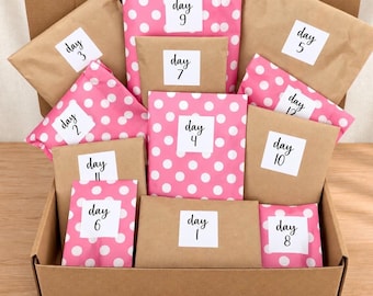 Op de afbeelding: Een kartonnen doos gevuld met twaalf individuele pakketjes verpakt in bruin papier of roze papier met witte stippen. Elk pakketje is voorzien van een wit vierkant label met zwarte tekst die een dag van één tot en met twaalf aangeeft.