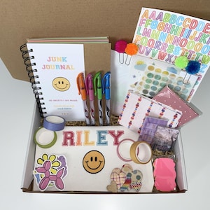 Può includere: Una scatola piena di articoli di cancelleria, tra cui un taccuino "Junk Journal" a spirale, penne colorate, adesivi, nastro washi e fermagli decorativi. La scatola contiene una varietà di articoli con un'estetica allegra e colorata.