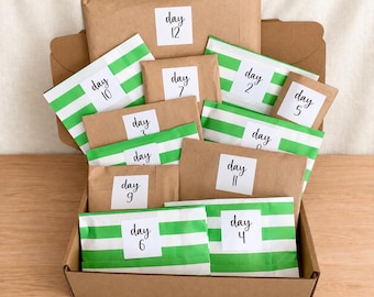Op de afbeelding: Een kartonnen doos gevuld met twaalf individuele pakketjes, sommige van bruin papier en andere met groene en witte horizontale strepen. Elk pakketje heeft een wit vierkant label met zwarte tekst van day 2 tot en met day 12.