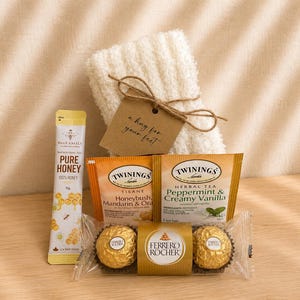 Puede incluir: Un set de regalo que incluye un par de calcetines blancos afelpados con una etiqueta marr&oacute;n que dice a hug for your feet, un palito de miel BeeFamily, dos bolsitas de t&eacute; Twinings y dos bombones de chocolate Ferrero Rocher.