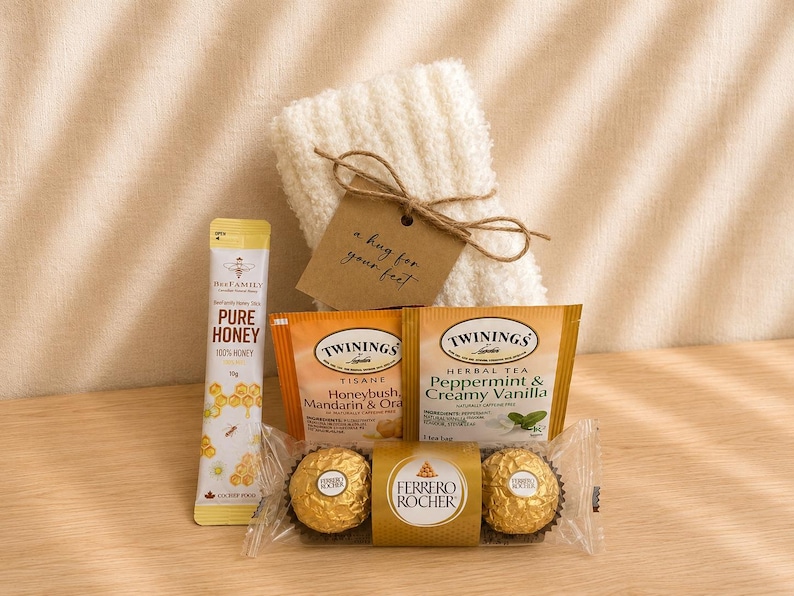 Puede incluir: Un set de regalo que incluye un par de calcetines blancos afelpados con una etiqueta marr&oacute;n que dice a hug for your feet, un palito de miel BeeFamily, dos bolsitas de t&eacute; Twinings y dos bombones de chocolate Ferrero Rocher.