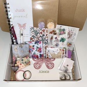Op de afbeelding: Een knutseldoos gevuld met journaling-benodigdheden. Inclusief een spiraalgebonden notitieboek met de woorden "Junk Journal", pennen, stickers, decoratief papier, washitape en versieringen. De doos is versierd met vlinder- en bloemmotieven.