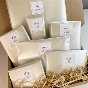 May include: A gift box containing seven wrapped presents, each marked with a day of the week. The gifts are wrapped in cream-coloured paper and have square labels. The box is filled with shredded paper for protection.