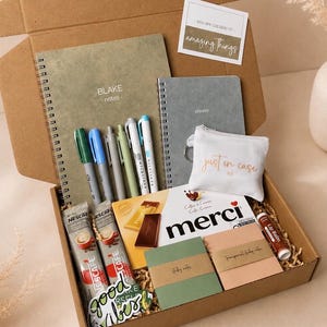 May include: A gift box filled with stationery and self-care items. Includes notebooks, pens, a small pouch, lip balm, coffee packets, and chocolate. The box contains the text "BLAKE notes", "planner", "just in case", and "good vibes".