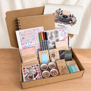 May include: A craft supply kit in a cardboard box containing a spiral-bound notebook, a zipped pouch with a typewriter illustration labelled Coraline, alphabet stickers, four Staedtler pens, handmade envelopes, wooden embellishments, and ribbons.