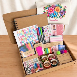 Puede incluir: Un kit de manualidades en una caja de cartón que contiene un cuaderno de espiral, una hoja de pegatinas del alfabeto coloridas, tres rotuladores, sobres hechos a mano, una herramienta rosa, cinta washi, tarjetas y un estuche floral.