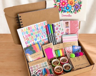 May include: A craft supply kit in a cardboard box containing a spiral-bound notebook, a sheet of colorful alphabet stickers, three markers, patterned envelopes, washi tape, journal cards, pom-pom page savers, and a floral-printed canvas pouch.