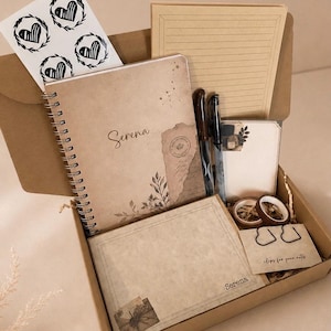 May include: A stationery gift box containing a spiral-bound notebook with the name Serena, a lined notepad, a small notepad, two pens, two rolls of washi tape, a sheet of heart stickers, and two heart-shaped paper clips on a card labeled clips for your notes.