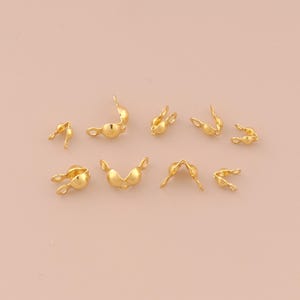 Könnte beinhalten: Eine Sammlung kleiner, goldfarbener Schmuckzubehörteile, darunter Quetschperlen und andere Komponenten. Diese Teile sind für die Schmuckherstellung konzipiert und haben eine glänzende, metallische Oberfläche. Die Fundstücke sind auf einer hellrosa Oberfläche verstreut.