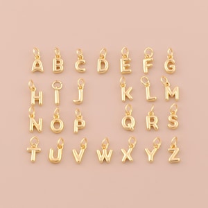 May include: Gold-colored alphabet charms, each with a small loop for attaching to jewelry. The letters A through Z are displayed, all in a simple, elegant font. The charms are arranged on a light pink background.