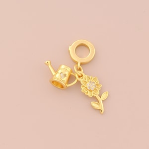 Puede incluir: Un charm dorado con un diseño de regadera con el texto "unfold & bloom" y un girasol con un cristal central. Adecuado para joyería o proyectos de manualidades.