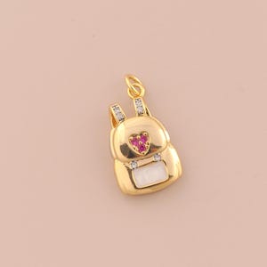 May include: A gold-colored backpack charm with a heart-shaped design in the center, adorned with small pink gems. The charm has a white rectangular detail at the bottom and is embellished with clear crystals.