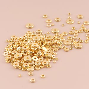 Puede incluir: Un montón grande de cuentas espaciadoras de metal de color dorado. Las cuentas son redondas con un agujero en el centro. Las cuentas están dispersas sobre una superficie rosa claro.