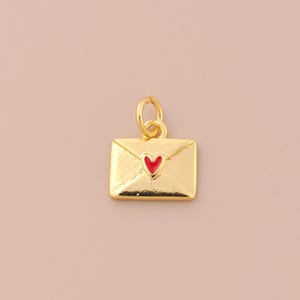 May include: A gold-colored pendant in the shape of an envelope with a red heart in the center. The pendant has a small loop at the top for attaching to a chain or bracelet. The background is a solid, light pink color.