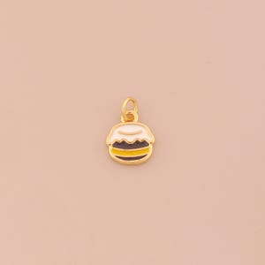 Puede incluir: Un dije dorado con forma de postre en capas, posiblemente un macaron o un pastel similar. El dije presenta detalles de esmalte blanco, marrón y amarillo, con un lazo dorado para sujetarlo a un collar o pulsera. El fondo es de color melocotón claro.