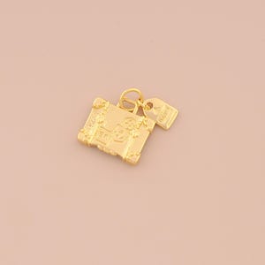 May include: A gold-colored charm in the shape of a vintage suitcase with a matching luggage tag. The suitcase charm has intricate embossed details, including a central emblem and corner accents. The luggage tag is rectangular with embossed text.