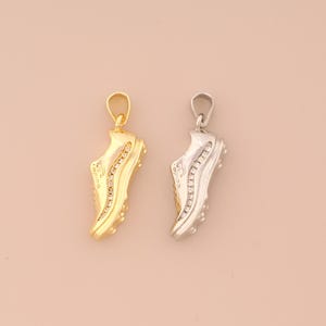 May include: Two pendants shaped like running shoes. One is gold-colored with clear stones, the other is silver-colored with clear stones. Both have a loop at the top for a chain. The background is a solid, light peach color.