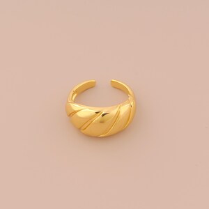 Puede incluir: Un anillo de color dorado con un diseño texturizado y curvo. El anillo es abierto y ajustable.