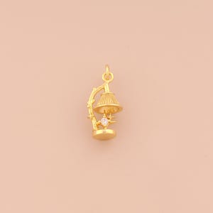May include: A gold-colored charm in the shape of a small lamp. The lamp has a detailed lampshade, a decorative base, and a clear gemstone. The charm is suspended from a loop at the top, suggesting it's designed to be worn as a pendant.