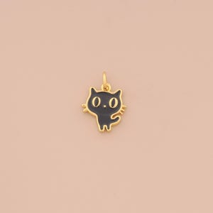 May include: A gold-colored cat charm with black enamel detailing. The charm features a stylized cat design with large eyes and a small tail. The charm is suspended from a small loop, ready to be added to a necklace or bracelet.
