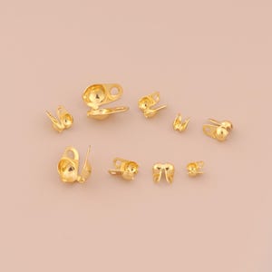 May include: A collection of small, gold-colored metal clasps and findings, including hook and eye closures and decorative elements. The clasps vary in size and design, with some featuring a rounded shape and others with a more angular appearance. These findings are likely used in jewelry making or garment construction.