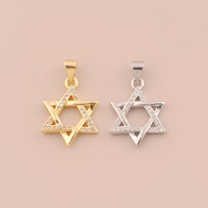 May include: Two Star of David pendants. One is gold-colored, the other is silver-colored. Both pendants feature a six-pointed star design with small, clear stones set along the edges. The pendants are displayed against a light pink background.