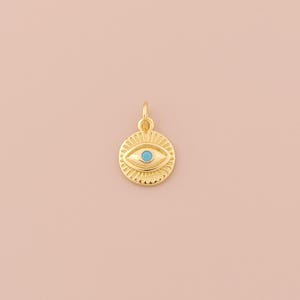 Könnte beinhalten: Goldener Anhänger mit einem blauen bösen Auge. Der Anhänger ist rund und hat eine strukturierte Oberfläche.