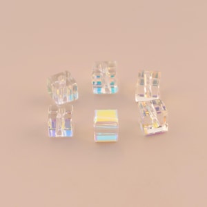 May include: Six clear, iridescent cube-shaped beads. Each bead has a hole running through the center. The beads reflect a rainbow of colors, including yellow, blue, and purple. The beads are scattered on a light pink surface.