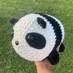 May include: A handmade, plush panda bear with a round body. The panda has a white body with black accents, including the ears, eyes, and a stripe across the back. The eyes are large and black. The panda is made of soft, textured material.