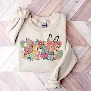 May include: A beige sweatshirt with a graphic design of the word "NURSE" in colorful letters surrounded by flowers and Easter eggs. The graphic also includes a bunny rabbit with a bow.