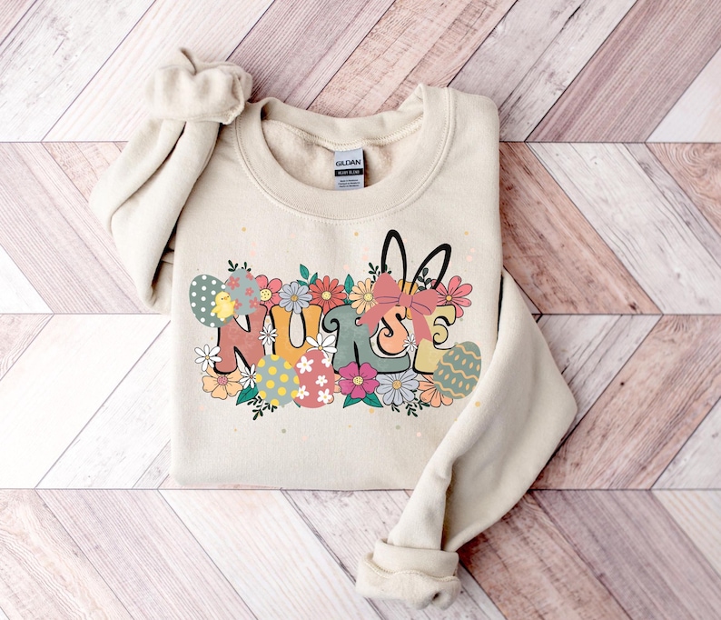 May include: A beige sweatshirt with a graphic design of the word "NURSE" in colorful letters surrounded by flowers and Easter eggs. The graphic also includes a bunny rabbit with a bow.