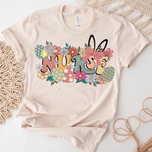 May include: A beige t-shirt with a graphic design of the word "NURSE" surrounded by colorful flowers and Easter eggs. The graphic also includes a bunny rabbit with a pink bow.