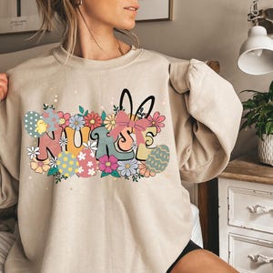 May include: A beige sweatshirt with a graphic design of the word "NURSE" with Easter eggs and flowers. The graphic is in pastel colors.